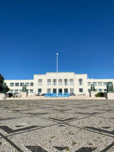 Entrance to the white Tecnico Lisboa Bulding in front of a bluke sky. in Front of the building are blue blocks saying @tecnico