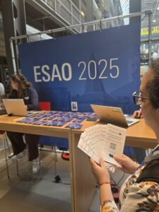 BioMembrOS leaflets fanned out in a woman's hands. Behind her is a wooden table with a blue ESAO 2025 banner with white lettering hanging behind it.