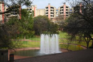 A fountain inside a little pond, surrounded by trees and lawn. In the background a building of the university of Johannesburg