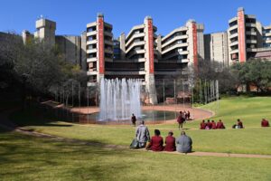 Fountain in the middle, surrounded by trees and lawn with Students in red and grey uniforms sitting on it. In the back a building of the university of Johannesburg